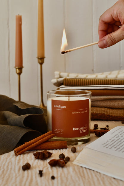 Cardigan candle next to books, spices and a lit match, front view.